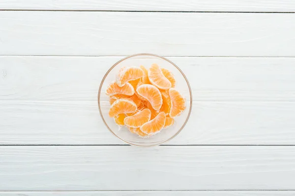 Вид сверху на очищенные ломтики мандарина в стеклянной чаше на деревянном белом фоне — стоковое фото