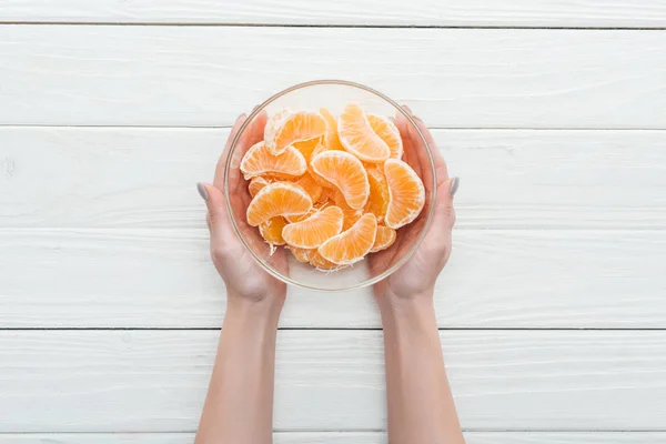 Vista ritagliata di donna che tiene ciotola di vetro con fette di mandarino su sfondo bianco in legno — Foto stock