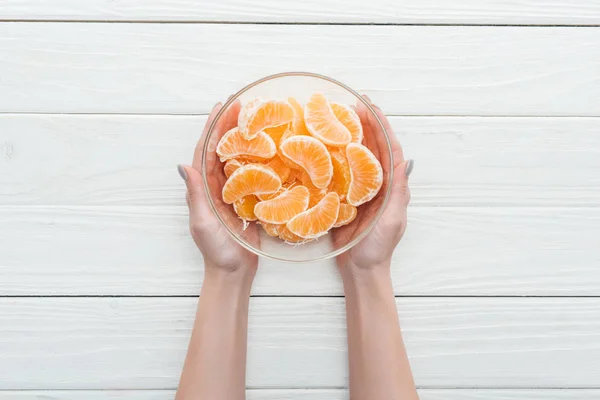 Vista recortada de la mujer sosteniendo tazón de vidrio con rodajas de mandarina sobre fondo blanco de madera - foto de stock