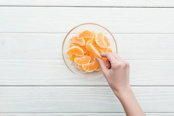 Vista cortada da mulher tomando fatias de tangerina de tigela de vidro no fundo branco de madeira — Fotografia de Stock