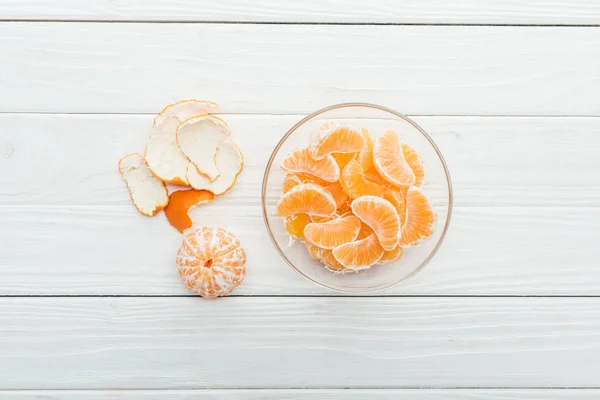 Top view of peeled tangerine slices in glass bowl and peel on wooden white background — Stock Photo