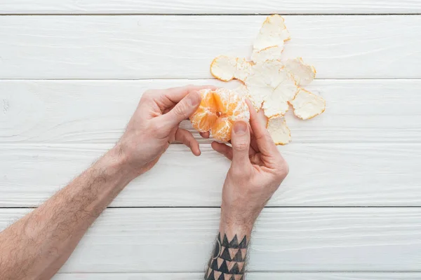 Partial view of tattooed man holding peeled tangerine on white wooden surface — Stock Photo