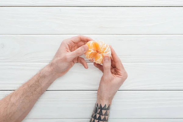 Cropped view of tattooed man holding peeled tangerine on white wooden surface — Stock Photo