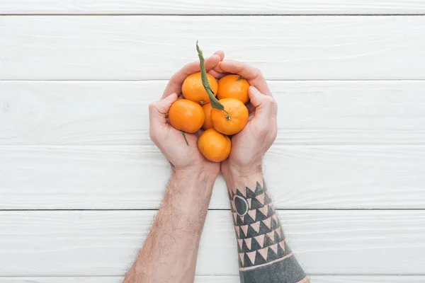 Cropped view of tattooed man holding heap of tangerines on white wooden surface — Stock Photo