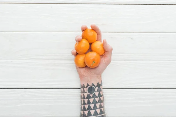 Cropped view of tattooed man holding heap of tangerines in hand on white wooden surface — Stock Photo