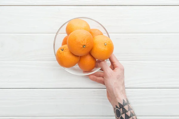 Cropped view of man holding glass bowl with ripe tangerines on wooden surface — Stock Photo