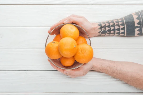 Vue recadrée d'un homme tatoué tenant un bol en verre avec des mandarines mûres sur une surface en bois — Photo de stock