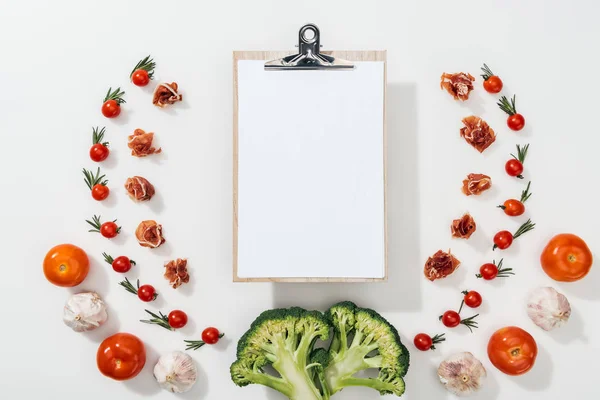 Top view of empty clipboard among tomatoes, leaves, broccoli, prosciutto and garlic — Stock Photo