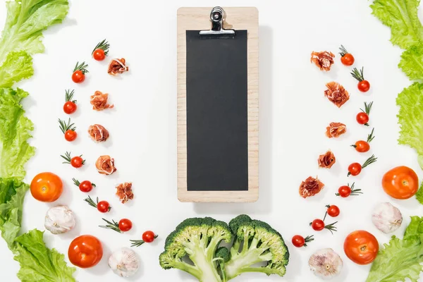 Top view of clipboard among tomatoes, lettuce leaves, prosciutto, broccoli and garlic — Stock Photo