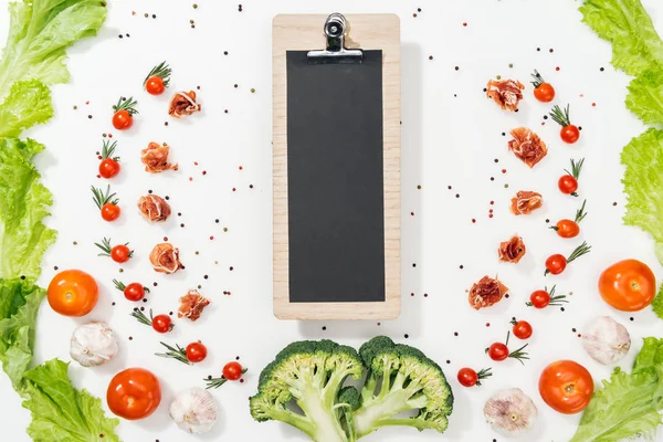 Top view of black clipboard among tomatoes, lettuce leaves, prosciutto, broccoli, spices and garlic — Stock Photo