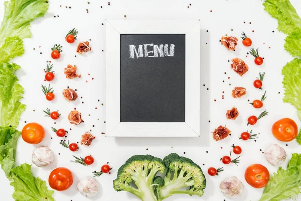 Chalk board with menu lettering among tomatoes, lettuce leaves, prosciutto, broccoli, spices and garlic — Stock Photo