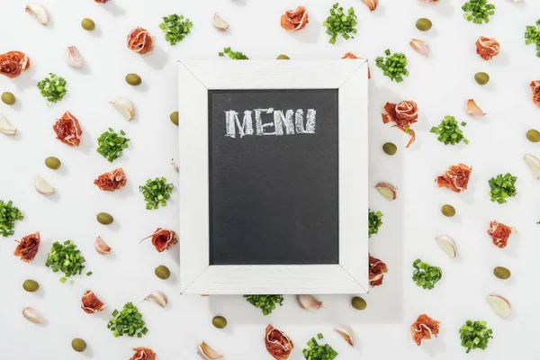 Vista dall'alto su lavagna di gesso con menù lettering tra prosciutto, olive, spicchi d'aglio e verde — Foto stock