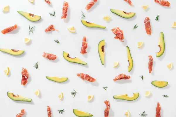 Vue du haut du prosciutto près des avocats tranchés, citrons et brindilles de romarin sur fond blanc — Photo de stock