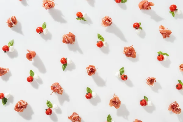 Vista superior de prosciutto saboroso perto de tomates cereja vermelhos, queijo mussarela e folhas de manjericão no fundo branco — Fotografia de Stock