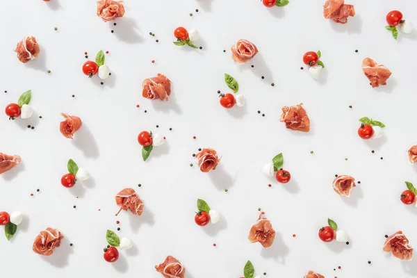 Top view of tasty prosciutto near red cherry tomatoes, mozzarella cheese and peppercorns on white background — Stock Photo