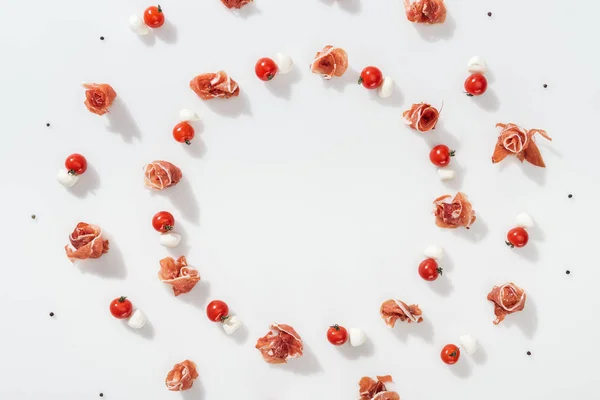 Flat lay of tasty prosciutto near red cherry tomatoes, mozzarella cheese and peppercorns on white background — Stock Photo