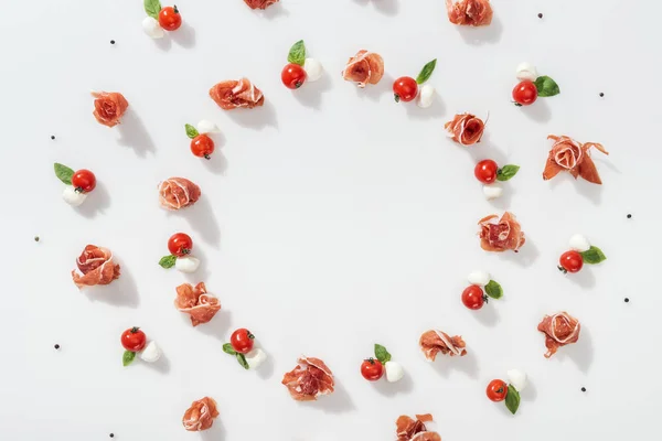 Puesta plana de sabroso prosciutto cerca de tomates rojos cereza, queso mozzarella y granos de pimienta sobre fondo blanco - foto de stock