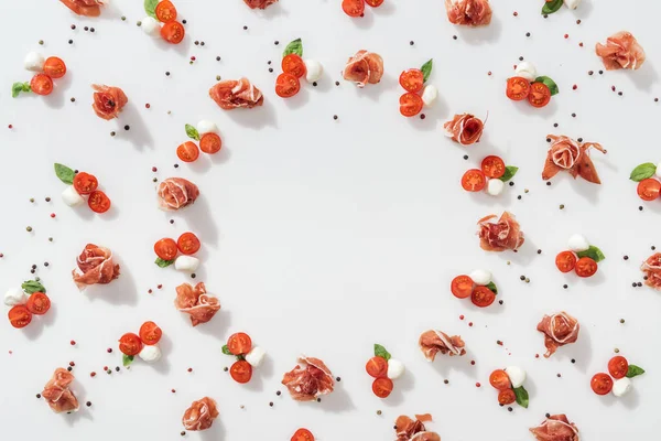 Puesta plana de jamón cerca de tomates rojos cereza, queso mozzarella, hojas de albahaca verde y granos de pimienta sobre fondo blanco - foto de stock