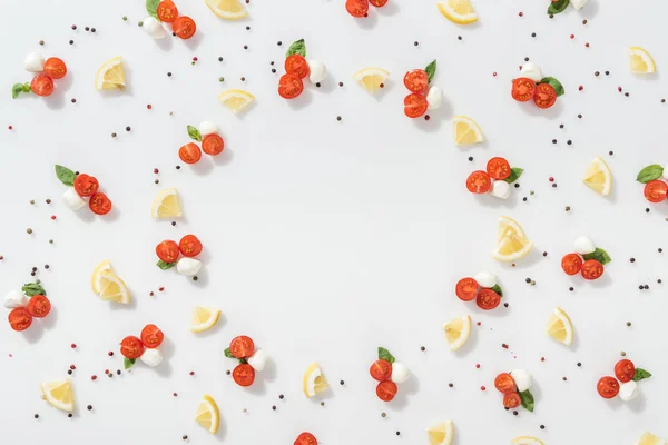 Puesta plana de tomates rojos cereza, queso mozzarella, hojas de albahaca verde y rodajas de limones cerca de granos de pimienta sobre fondo blanco - foto de stock