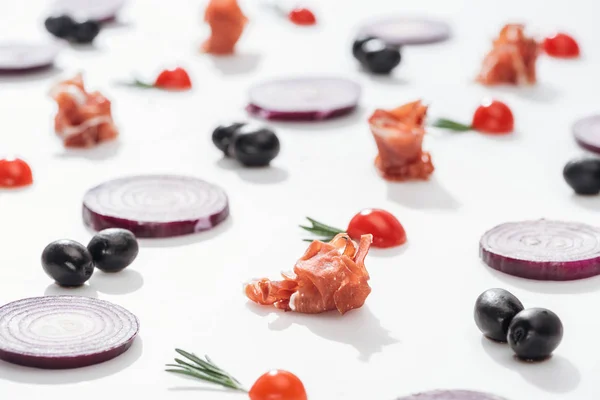 Enfoque selectivo de jamón crudo cerca de tomates cherry con ramitas de romero cerca de anillos de cebolla roja y aceitunas negras sobre fondo blanco - foto de stock
