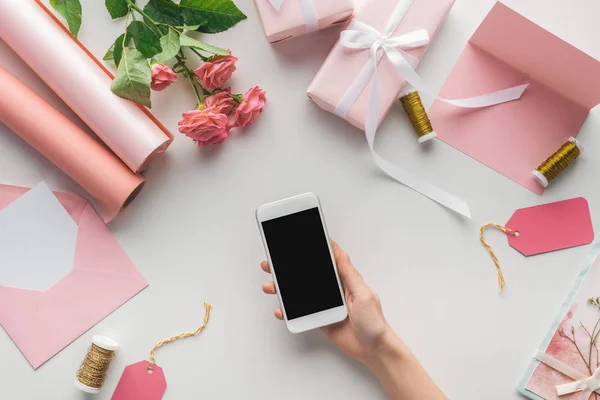 Vue recadrée d'une femme tenant un smartphone près de roses, des rouleaux de papier, des cadeaux emballés, une enveloppe, une carte de vœux et des bobines de fil sur fond gris — Photo de stock