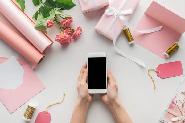 Vista ritagliata di donna che tiene smartphone vicino a rose, rotoli di carta, regali avvolti, busta, carta e bobine di filo su sfondo grigio — Foto stock