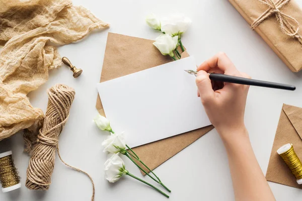 Vista recortada de la mujer escribiendo en tarjeta con pluma de tinta cerca de eustoma, tela, sobre y regalo envuelto en fondo gris - foto de stock