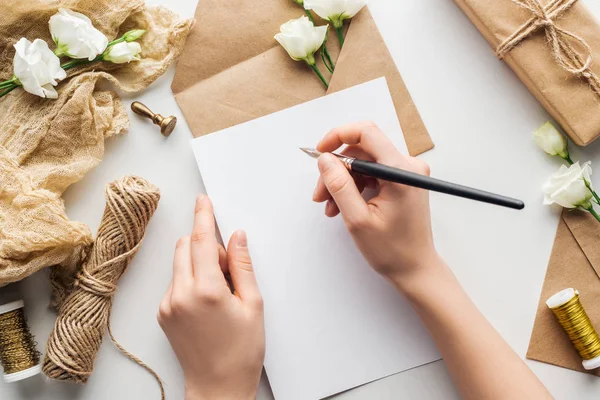 Vista ritagliata di donna che scrive in carta con penna inchiostro vicino a fiori, stoffa, busta e regalo avvolto su sfondo grigio — Foto stock