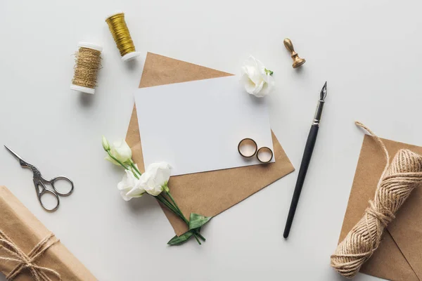 Top view of envelope with empty card, ink pen, flowers, stamp, wrapped gift, scissors, spools of thread and golden wedding rings on grey background — Stock Photo
