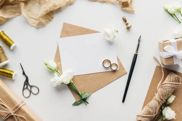Top view of empty card with envelope, ink pen, eustoma, stamp, wrapped gifts, scissors, twine and golden wedding rings on grey background — Stock Photo