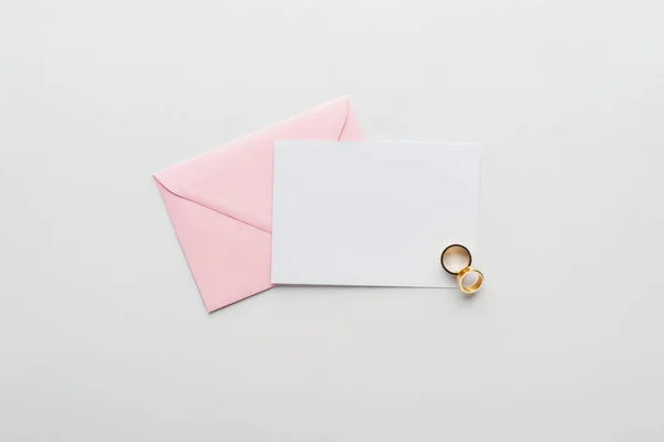 Вид сверху на пустую открытку с розовым конвертом и золотыми обручальными кольцами на сером фоне — стоковое фото