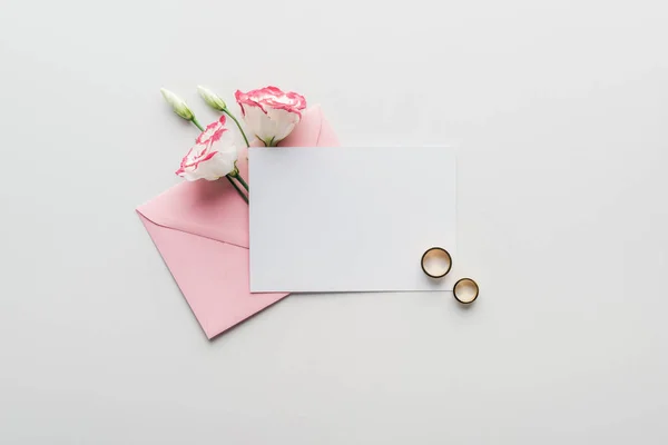 Вид сверху на пустую открытку с розовым конвертом, цветы и золотые обручальные кольца на сером фоне — стоковое фото