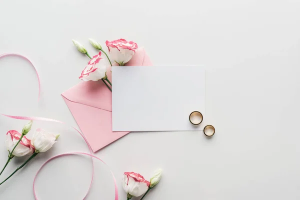 Top view of empty card with pink envelope, flowers, silk ribbon and golden wedding rings on grey background — Stock Photo