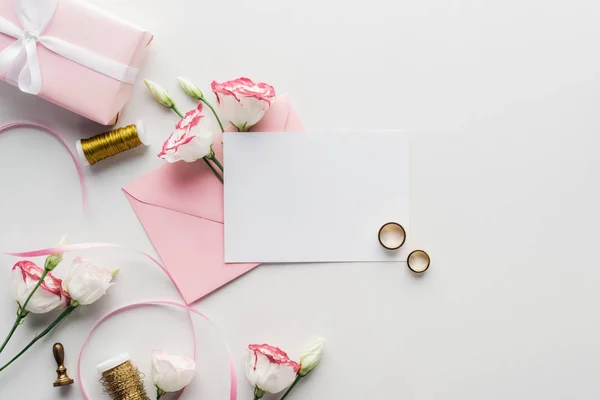 Вид сверху на пустую открытку с розовым конвертом, цветы, шелковая лента, завернутый подарок, печать и золотые обручальные кольца на сером фоне — стоковое фото