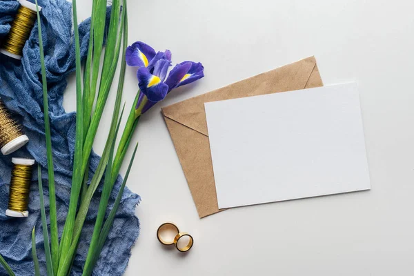Vista superior de la tarjeta vacía con sobre, flores, tela azul, carretes de hilo y anillos de boda dorados sobre fondo gris - foto de stock