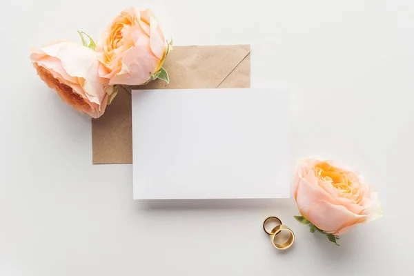 Top view of empty card over brown craft paper envelope, roses and golden wedding rings on grey background — Stock Photo
