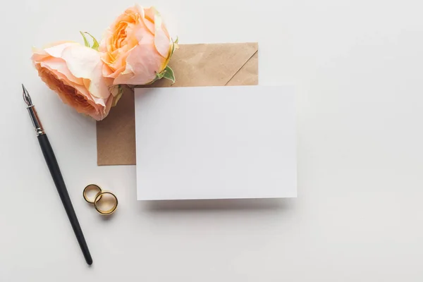 Vista superior do cartão vazio sobre envelope de papel de ofício marrom, caneta de tinta, rosas e anéis de casamento dourados no fundo cinza — Fotografia de Stock