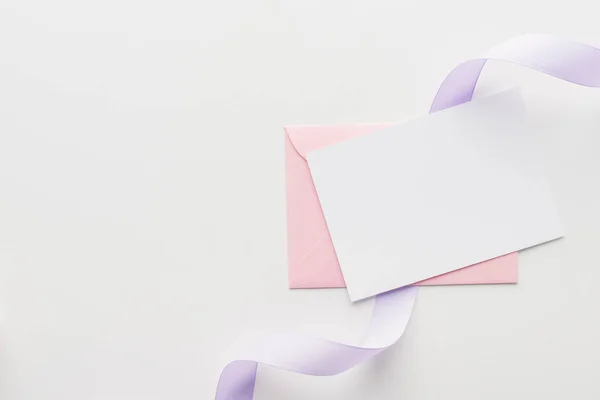 Вид сверху на пустой лист с розовым конвертом и фиолетовой шелковой лентой на сером фоне — стоковое фото