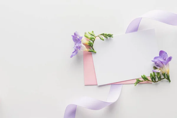 Vista superior de blanco vacío con sobre rosa, flores púrpura y cinta de seda sobre fondo gris - foto de stock