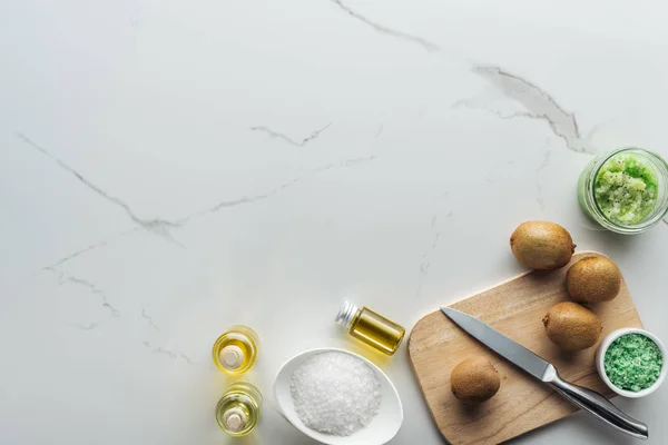 Draufsicht auf Schalen und Gefäße mit hausgemachten kosmetischen Zutaten, Holzschneidetisch mit Messer und Kiwi auf weißer Oberfläche — Stockfoto