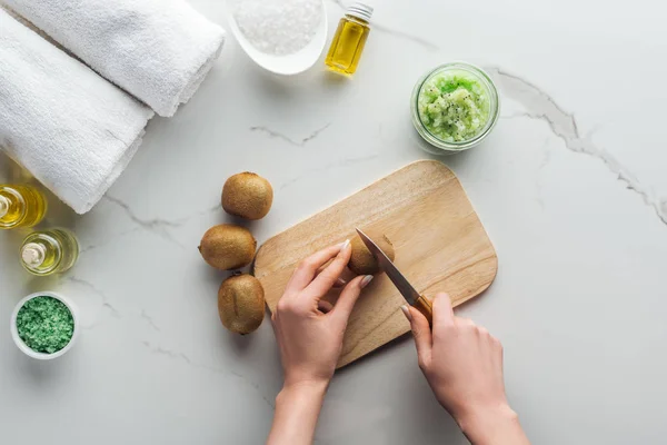 Teilansicht einer Frau, die Kiwi auf einem hölzernen Schneidetisch schneidet, und verschiedene natürliche Zutaten für handgefertigte Kosmetik auf weißer Oberfläche — Stockfoto