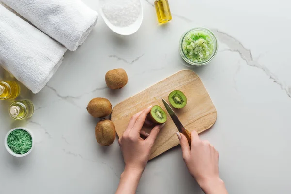 Partial view of woman cutting kiwi on wooden cutting desk, and different ingredients for making cosmetics on white surface — Stock Photo