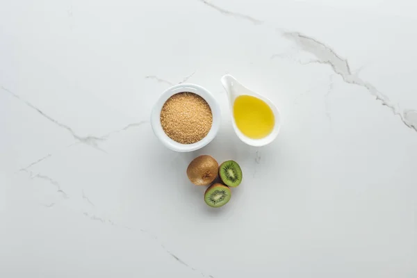 Vista superior de kiwi entero y cortado, tazones con azúcar morena y aceite en la superficie blanca - foto de stock