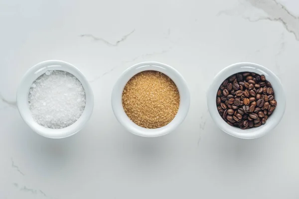 Vista dall'alto di ciotole con sale, chicchi di caffè e zucchero di canna sulla superficie bianca — Foto stock