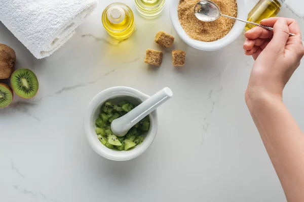 Cropped view of woman holding spoon of brown sugar, pounder with kiwi, and various cosmetic ingredients on white surface — Stock Photo