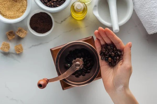 Vue partielle de la main féminine avec grains de café, moulin à main, pounder et différents ingrédients pour la fabrication de cosmétiques sur surface blanche — Photo de stock