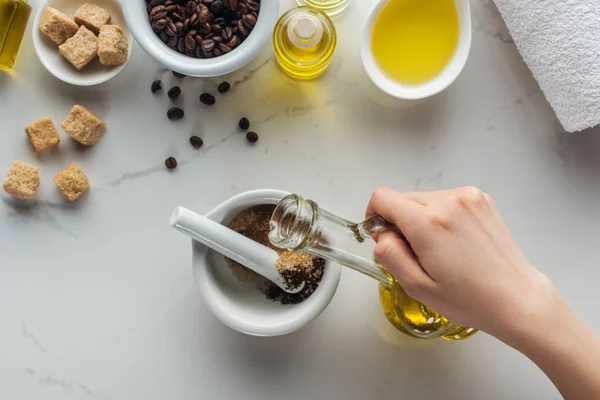 Cropped view of woman adding oil into pounder with bround sugar and ground coffee on white surface — Stock Photo