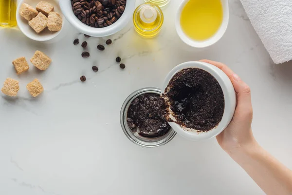 Vista recortada de la mujer vertiendo exfoliante de café casero en el recipiente de vidrio en la superficie blanca - foto de stock