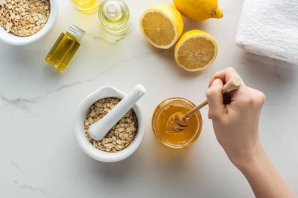 Visão parcial da mão feminina com vara de mel, pounder com flocos de aveia, limões e diferentes ingredientes naturais na superfície branca — Fotografia de Stock