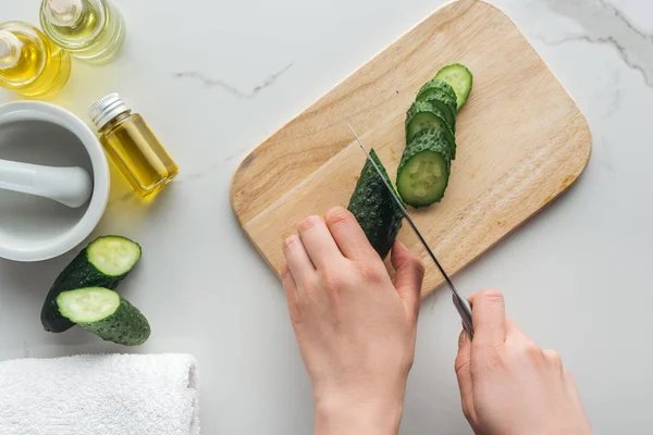 Cropped view of woman cutting cucumber on wooden cutting desk, pounder and various cosmetic ingredients on white surface — Stock Photo